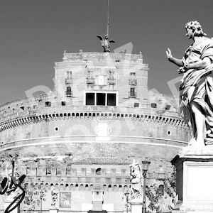 May include: Black and white photograph of the Castel Sant'Angelo in Rome, Italy. A large cylindrical building with a statue of an angel in the foreground. The sky is clear and the image is in grayscale.