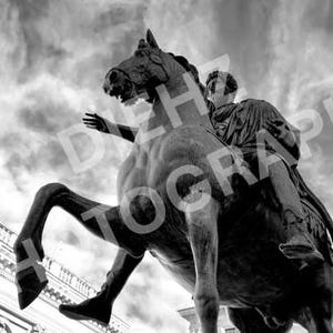 Puede incluir: Fotografía en blanco y negro de una estatua ecuestre de bronce contra un cielo nublado. La estatua representa una figura sobre un caballo encabritado, con el brazo del jinete extendido. Se ven edificios al fondo.