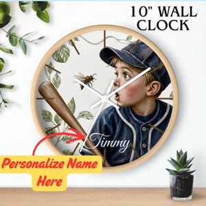 May include: A 10-inch round wall clock with a light wood frame. The clock face features an illustration of a child in a blue baseball uniform and cap holding a bat, with a bee and green leaves. The name Timmy is printed in white script.