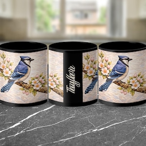 May include: Black ceramic mug featuring a blue jay perched on a branch with pink flowers. The mug has a black handle and interior. The name "Taylor" is printed on the mug in a script font.