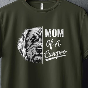 May include: Olive green t-shirt featuring a detailed black and white illustration of a Cavapoo dog's face. The text "MOM of A Cavapoo" is printed in white, with a distressed font. The shirt is displayed on a hanger.