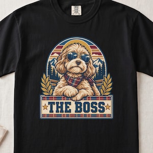 May include: A black t-shirt featuring a graphic of a light brown dog wearing sunglasses and a plaid bandana. The design includes a mountain landscape, wheat stalks, and a rectangular banner with the text THE BOSS in blue block letters.