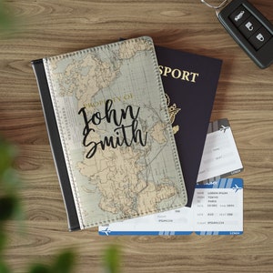May include: A passport holder with a vintage world map design. The holder is black with a stitched border and has the name "John Smith" printed in gold lettering. The holder is sitting on top of a blue passport and two airline boarding passes.