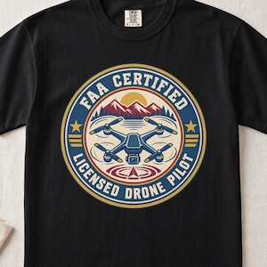 May include: A black t-shirt featuring a circular graphic with a drone in the center, mountains, and a sun. The text reads FAA CERTIFIED LICENSED DRONE PILOT. The design uses blue, red, yellow, and white colors with stars and stripes accents.