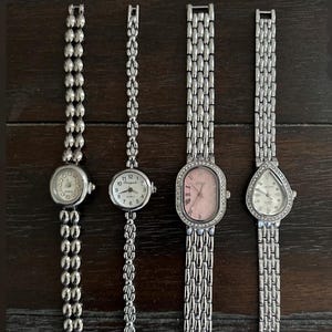 May include: Four silver wristwatches with varying band designs are shown. The watch faces are different shapes and colours, including round, oval, and teardrop. The bands feature unique patterns, such as beaded and linked styles.