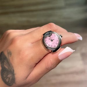 May include: A silver-toned ring watch with a pink face and black numerals. The watch has a heart-shaped face and a quartz movement. The ring is worn on a finger.