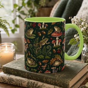 May include: A ceramic mug with a bright lime green interior and handle, featuring a dark green exterior surface decorated with a repeating pattern of frogs, red and white mushrooms, brown acorns, ferns, and autumn leaves in a forest theme.