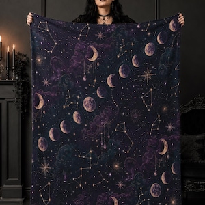May include: A rectangular throw blanket featuring a dark purple and black night sky pattern with various moon phases, glowing stars, and connected constellations. The design includes wispy clouds and hanging celestial charms.