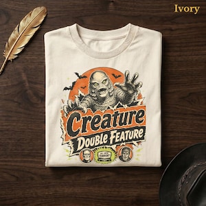 May include: An ivory t-shirt featuring a graphic print of a swamp creature emerging from an orange circle with bats. The text reads Creature Double Feature and All New Terror, with two small circular portraits of monster characters at the bottom.
