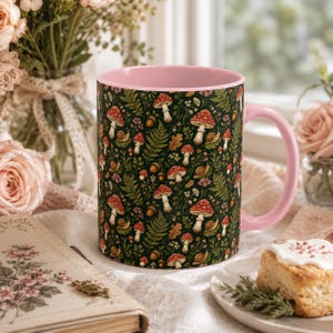 May include: A ceramic mug with a light pink interior and handle, featuring a dark green exterior surface decorated with a repeating pattern of red and white mushrooms, brown acorns, small snails, and green fern leaves.