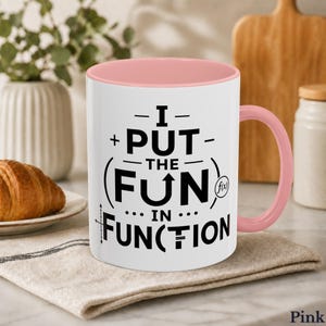 May include: White ceramic mug with a pink interior and handle. The mug features the text "I PUT THE FUN IN FUNCTION" in black, with a mathematical graph and the f(x) symbol. A croissant sits on a plate in the background.