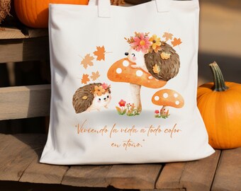 May include: White tote bag featuring an illustration of two hedgehogs with floral crowns and a mushroom. The design includes fall leaves and the text "Viviendo la vida a todo color en otoño." Pumpkins are in the background.