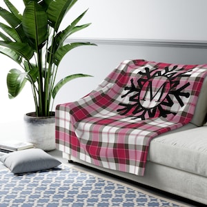 May include: A red, white, and gray plaid throw blanket with a black snowflake design and the letter "M" in the center. The blanket is draped over a gray sofa. A large green plant in a pot is in the background.