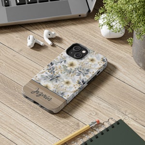 May include: A floral iPhone case with a light wood-grain bottom section. The case features a pattern of white and yellow flowers with gray and gold accents. The name "Jazmin" is printed on the wood-grain section.