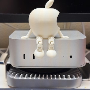 Pode incluir: Uma figura de maçã branca suja impressa em 3D, com braços e pernas, sentada em cima de um computador prateado. A maçã tem uma mordida e uma folha. O computador tem uma ventoinha e várias portas.