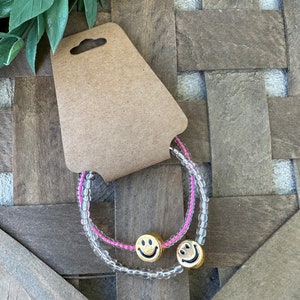 Op de afbeelding: Twee kralenarmbanden met smiley-bedels. De ene armband is helder met roze kralen en de andere is helder met witte kralen.