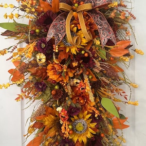 May include: A fall-themed door swag with a mix of orange, yellow, and burgundy flowers, leaves, and berries. A gold and burgundy ribbon with a paisley pattern is tied in a bow at the top.