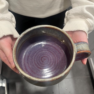 May include: A wide, round ceramic mug featuring a deep purple interior with visible concentric circular patterns. The exterior of the mug displays a mottled blend of earthy brown, grey, and blue tones with a thick, sturdy handle on the right side.