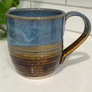 May include: A ceramic mug featuring a two-tone glaze with a mottled blue upper section and a dark brown, ribbed lower section. The mug has a curved handle and a smooth, rounded rim, showcasing a distinct transition between the two colors.