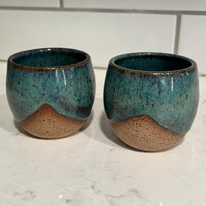 May include: Two small, rounded ceramic cups featuring a two-tone design. The upper portion is glazed in a speckled teal and dark blue, while the lower portion displays a natural, speckled tan clay finish with a curved, mountain-like border.