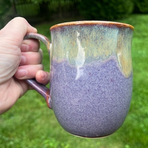 Peut inclure: Une tasse en céramique présentant un émail bicolore avec une base violette mouchetée et une partie supérieure lisse jaune-vert clair. Le bord et l'anse présentent une teinte brun-orangé chaude, créant un contraste net avec le corps de la tasse.