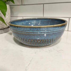 Peut inclure: Un bol en céramique bleu avec un extérieur texturé et un bord marron clair. Le bol a une forme ronde et est posé sur un comptoir blanc. Le bol est probablement utilisé pour servir des aliments ou comme pièce décorative.