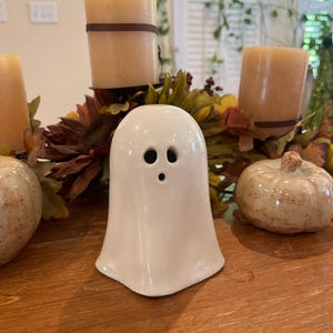 May include: A white ceramic ghost figurine with two black eye holes and a small mouth. The ghost is part of a fall-themed centerpiece with candles, pumpkins, and autumn foliage. The figurine is approximately 10 cm tall.