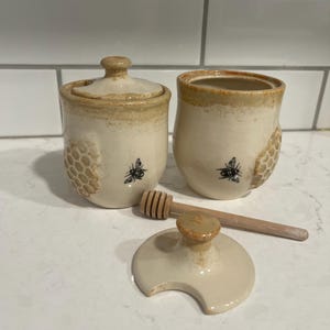 May include: A ceramic honey pot set with a lid, a honey dipper, and an open container. The set is off-white with a honeycomb pattern and a bee illustration. The lid is off the container, and the honey dipper is resting on the surface.