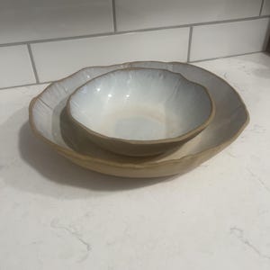 May include: Two ceramic bowls, one nested inside the other. The larger bowl has a wide, irregular rim and a light brown exterior. The interior of both bowls is white with a subtle, glossy finish, perfect for serving food.