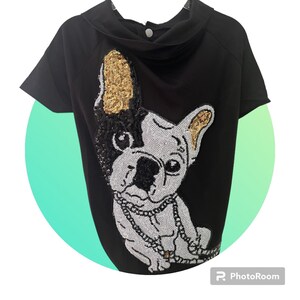 May include: Black short-sleeved top featuring a sequined French Bulldog design. The dog's face is white and black, with gold sequin accents on the ears. A silver sequin chain necklace adds detail to the design.