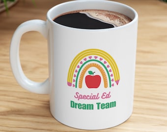 Può includere: Una tazza da caffè bianca con un design a forma di arcobaleno fatto di matite e cuori. L'arcobaleno ha una mela rossa al centro. Il testo "Special Ed Dream Team" è stampato sotto l'arcobaleno.