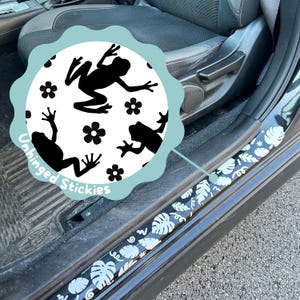 Pode incluir: Protetores de soleira de porta de carro em preto e branco com um padrão de folhas tropicais. Um adesivo circular com silhuetas de rãs pretas e flores também é visível. O adesivo tem o texto "Unhinged Stickies".