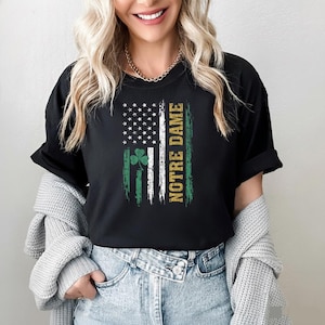 Può includere: Una t-shirt nera a maniche corte con una grafica invecchiata di una bandiera con stelle bianche, un trifoglio verde e il testo NOTRE DAME stampato verticalmente in oro. Il design incorpora strisce verticali nei colori verde, bianco e oro.