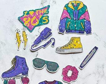 May include: A set of 10 colorful 80s themed stickers featuring a variety of items including a roller skate, a fanny pack, a sweater, a jacket, a hair straightener, a pair of sunglasses, a pair of earrings, a pair of sneakers, a hair scrunchie, and a toolbox. The stickers have a glittery finish.