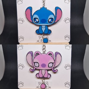 May include: Two Stitch character keychains, one blue and one pink, with a silver chain and a small charm at the bottom. The characters are cartoon-style and have large eyes and ears.