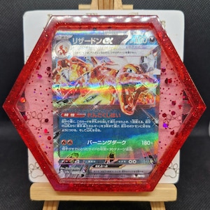 May include: A framed Pokémon trading card featuring Charizard EX. The card is encased in a red, hexagonal resin frame with glitter accents. The card art depicts a fiery dragon-like creature with Japanese text.