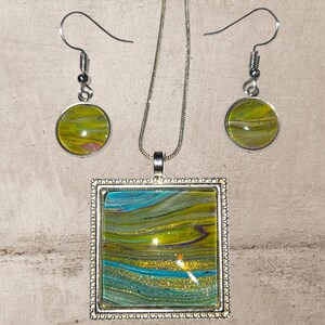 May include: A silver-toned jewelry set featuring a pendant necklace and matching earrings. The pendant is square, and the earrings are round. Each piece has a colorful abstract design with shades of blue, green, and gold. The set is displayed on a neutral background.