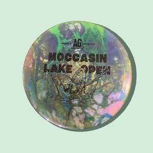May include: A disc golf disc with a vibrant, swirling design in shades of green, blue, and pink. The disc has the text "MOCCASIN LAKE OPEN" and a graphic of a disc creating a splash. Made by Above Gravity.