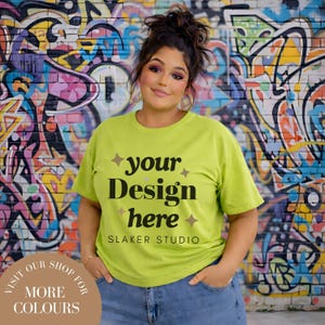 Può includere: Una maglietta verde lime con la scritta "your Design here" e "SLAKER STUDIO" in nero. La modella indossa jeans blu. Lo sfondo è un colorato muro di graffiti. Il testo "VISITA IL NOSTRO NEGOZIO PER PIÙ COLORI" è in un design circolare.