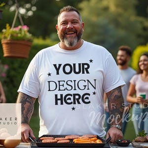 Puede incluir: Una camiseta blanca con el texto "YOUR DESIGN HERE" impreso en negro. La camiseta la lleva puesta una persona con barba y tatuajes. La persona está de pie frente a una parrilla con comida cocinada.