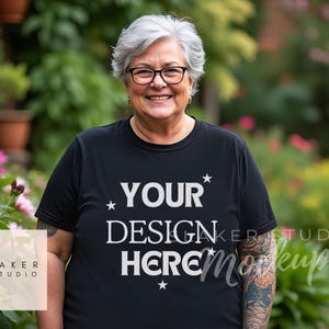 以下が含まれることがあります： 白い文字で「YOUR DESIGN HERE」とプリントされた黒いTシャツを着た人。緑の植物と花のある庭の前で立っている。