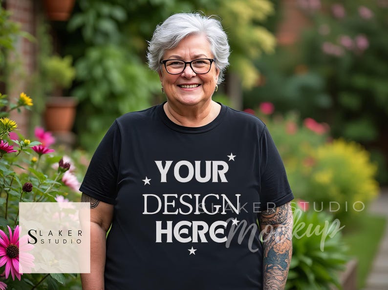 May include: A person wearing a black t-shirt with the text "YOUR DESIGN HERE" printed on it in white. The person is standing in front of a garden with green plants and flowers.