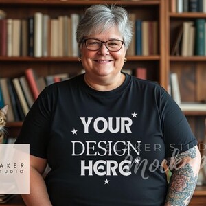 May include: A woman wearing a black t-shirt with the text "YOUR DESIGN HERE" printed on it. She is standing in front of a bookshelf with two cats sitting on the shelves below.