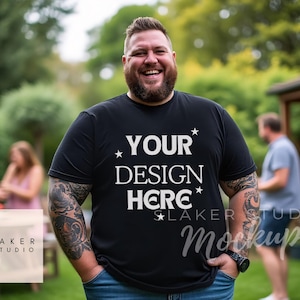 Puede incluir: Un hombre con una camiseta negra con el texto "YOUR DESIGN HERE" en blanco. Está de pie al aire libre con un césped verde y árboles en el fondo.