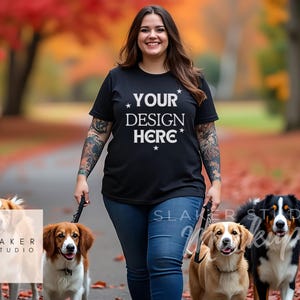 May include: A woman wearing a black t-shirt with the text "YOUR DESIGN HERE" walks with three dogs on leashes. The dogs are a brown and white dog, a brown and white dog, and a black and white dog. The woman is walking on a path with fallen leaves.