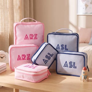 May include: A collection of rectangular packing cubes in pink and white or blue and white striped fabric. The pink bags feature the embroidered text ARZ, while the blue bags feature the embroidered text ASL. Each bag includes a top handle and zipper.