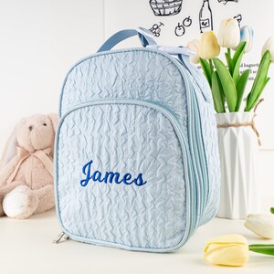 Pu&ograve; includere: Zaino imbottito azzurro chiaro con chiusura a zip e il nome "James" ricamato in blu sul davanti.