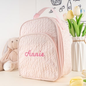 Pu&ograve; includere: Uno zaino rosa trapuntato con una tasca con cerniera e un nome ricamato rosa "Annie".