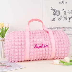 Op de afbeelding: Een roze, pluche, gewatteerde reistas met de naam "Sophia" geborduurd op de voorkant. De tas heeft twee handvatten en een ritssluiting.