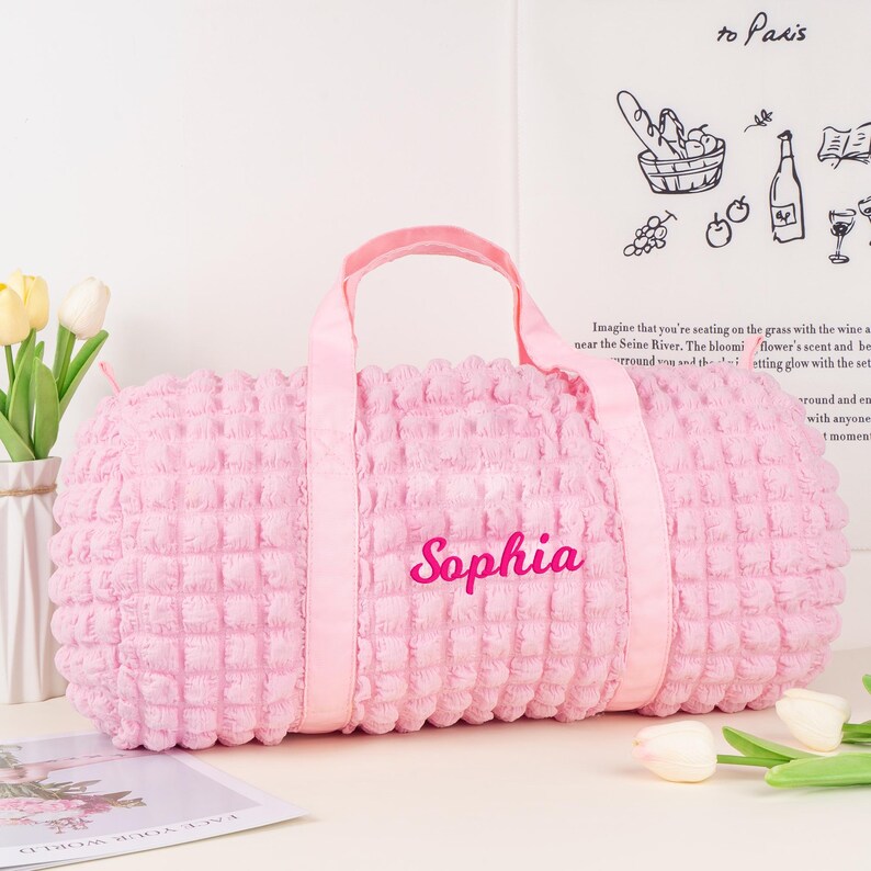Op de afbeelding: Een roze, pluche, gewatteerde reistas met de naam "Sophia" geborduurd op de voorkant. De tas heeft twee handvatten en een ritssluiting.